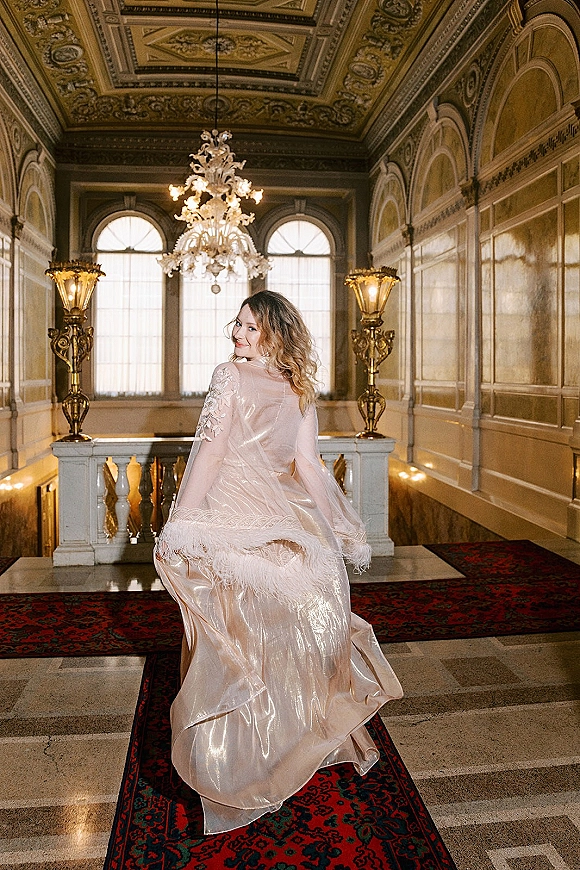 Bridal portrait of a bride looking back in an ornate ballroom, wearing a lace-sleeved gown with sheer overlay and chandelier above