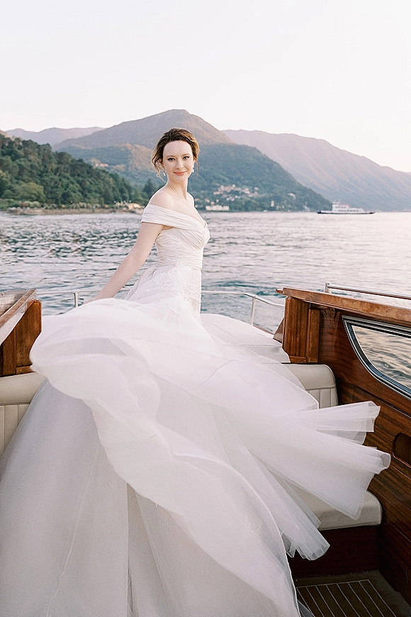 Bridal portrait of a bride in an off the shoulder wedding dress on boat, tulle skirt billowing on a wooden boat on a mountain lake