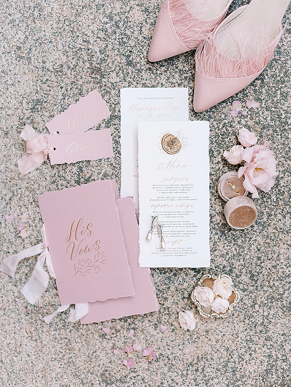 Invitation suite with blush wedding invitations, wax seal and deckled edges, rings, pearl earrings and pink heels on stone with petals