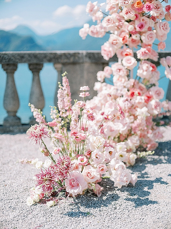 Wedding floral installation with a pink rose arch and blush blooms, with ground arrangements by a stone balustrade and mountain view