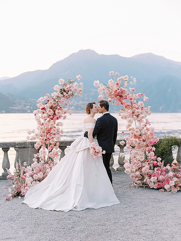 Wedding kiss portrait of bride and groom kissing under a pink floral arch, with lake and mountains behind on a stone terrace
