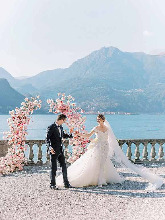 Couple portrait of bride and groom holding hands beneath a pink floral arch on a stone terrace with lake and mountain views behind