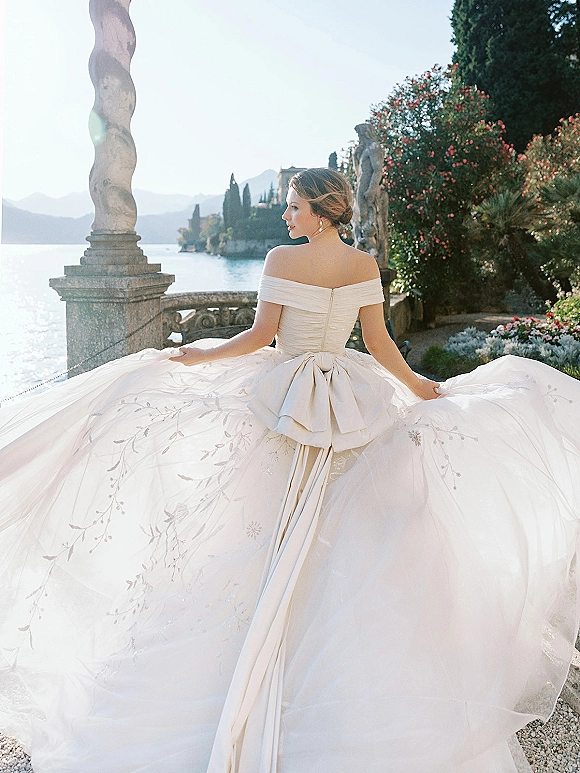 Bridal portrait of a bride in an off the shoulder wedding dress with an oversized bow, standing on a lakeside terrace with mountains beyond