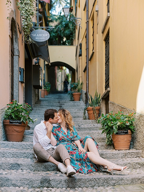 Engagement couple portrait of a couple kissing on stone steps, she in a blue floral wrap dress, in a narrow ivy-lined alley archway