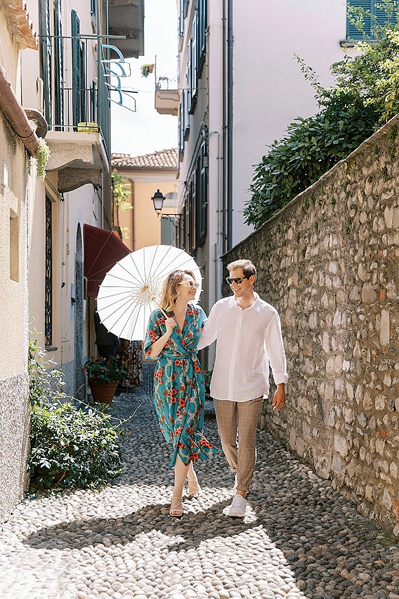 Couple portrait walking with a white parasol and sunglasses, she in a floral dress beside white buildings on a cobblestone street