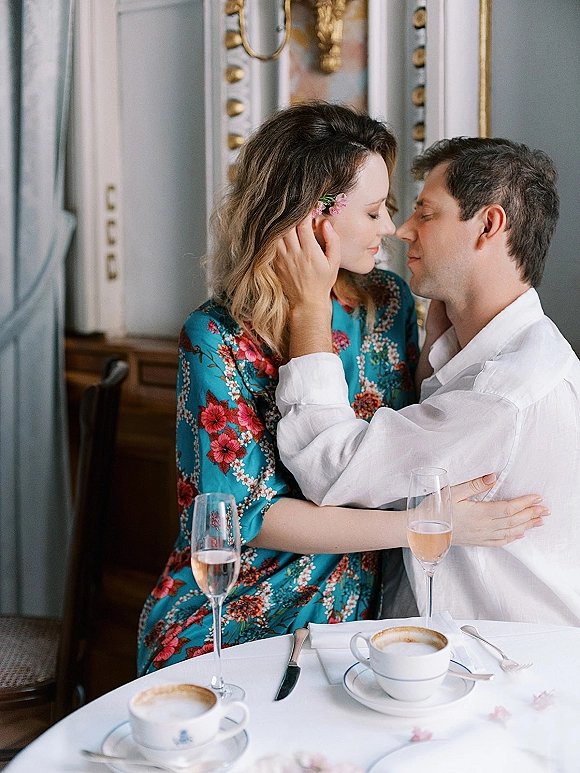 Couple portrait with forehead touch, seated at a cafe table with champagne flutes, coffee cups, and a mirror backdrop behind them