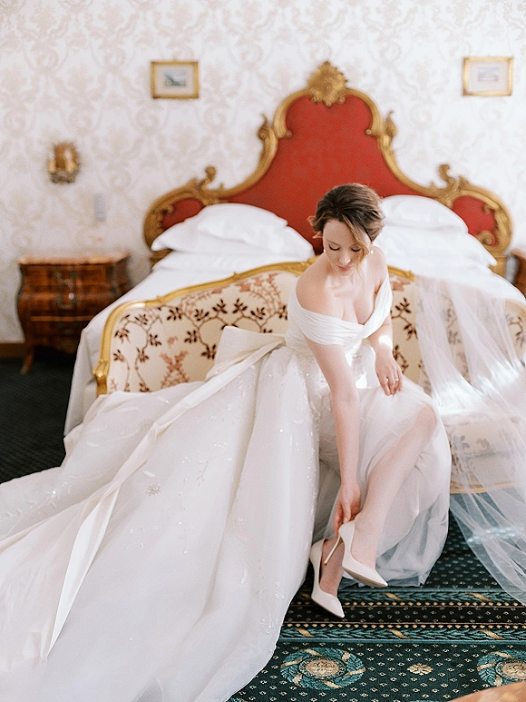 Bridal portrait of a bride putting on heels in an off the shoulder wedding dress with cathedral veil in a vintage hotel suite