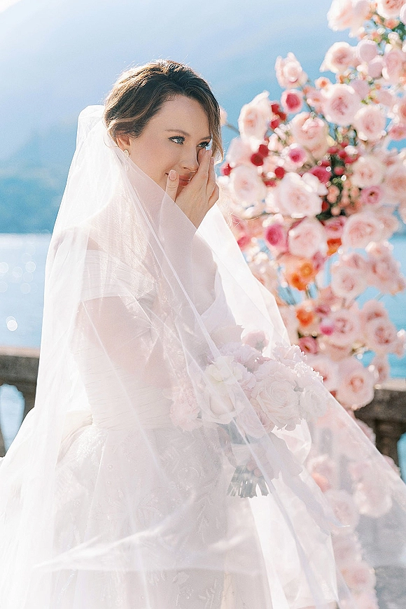 Bridal portrait of a bride with veil holding a soft pink rose bouquet, earrings and gown framed by a floral arch above ocean and stone railing