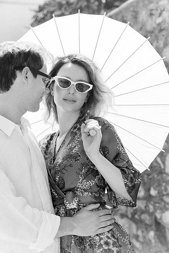 Couple portrait in outdoor greenery, embracing under a paper parasol; bride in cat-eye sunglasses and floral dress shows ring