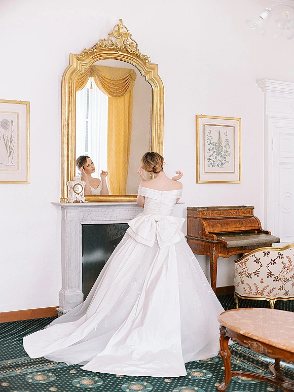 Bridal portrait of a bride looking in mirror, wearing an off-the-shoulder gown with oversized bow and long train in a vintage sitting room