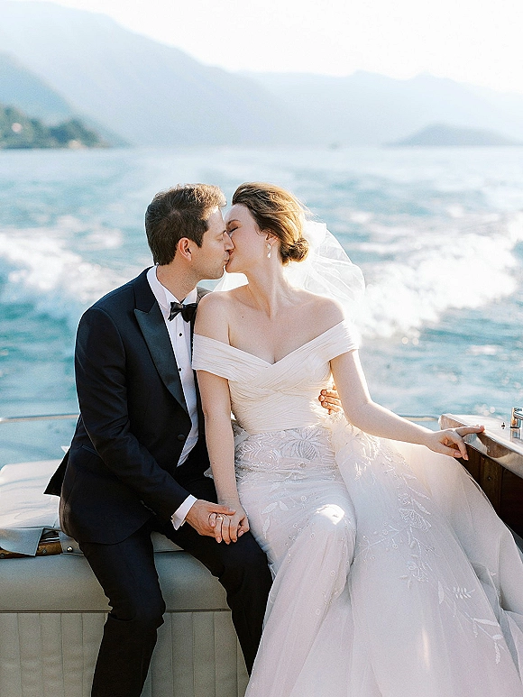 Wedding kiss portrait of bride and groom kissing on a boat deck, her veil blowing, with mountain lake and water wake behind them
