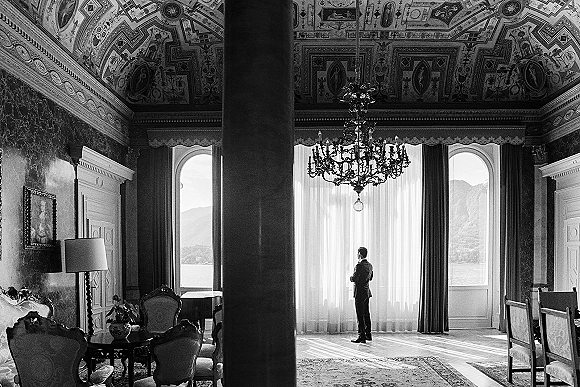 Groom portrait in a suit by an arched window, gazing out over a lake and mountains in a chandelier-lit, elegant suite