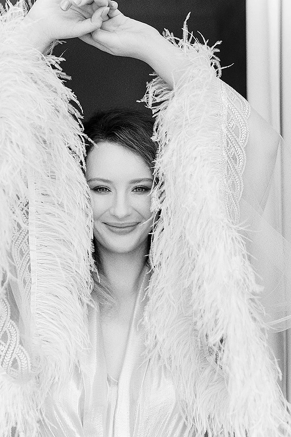 Bridal portrait of a bride in a feather robe with lace sleeves and a sheer veil, softly lit by window light against an indoor wall