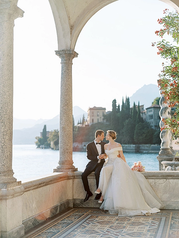 Couple portrait of bride and groom sitting and holding hands on a stone terrace with arched columns, lake and mountains behind, bouquet accent