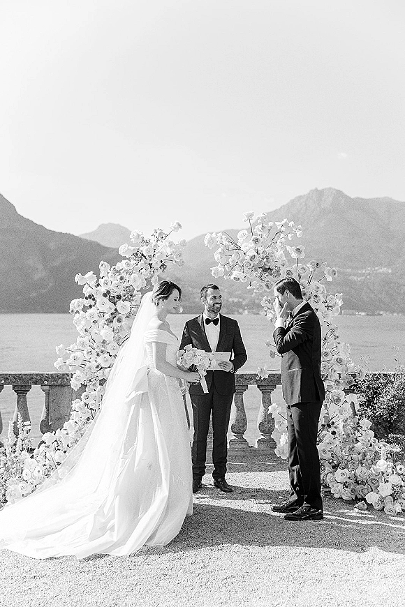 Wedding ceremony moment as bride and groom stand under a white rose floral arch on a stone terrace by a mountain lake, long veil flowing