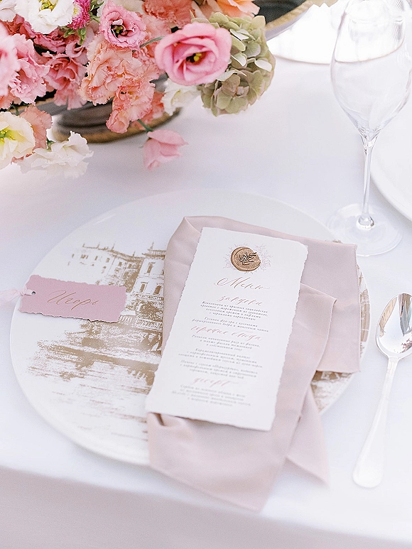 Wedding place setting with wedding menu card sealed in wax, blush napkin, pink place card, and floral centerpiece on white tablecloth