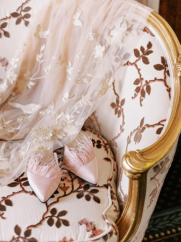 Bridal shoes in blush wedding heels with feather trim resting on a gold upholstered chair, veil draped over embroidered fabric and floral upholstery