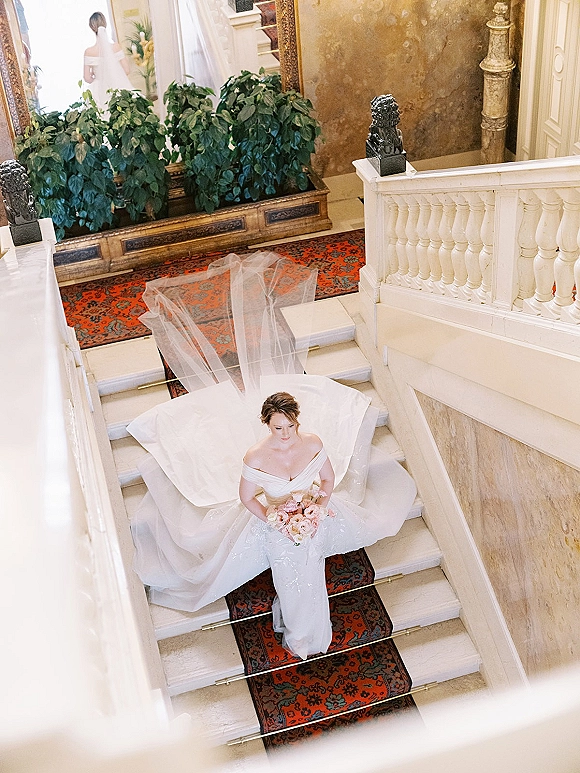 Bridal portrait of a bride on staircase, her off-the-shoulder gown and cathedral veil flowing beside a pastel bouquet in a grand hall