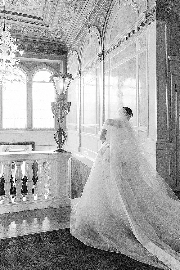 Bridal portrait of a bride from behind in an off the shoulder gown with long veil and train on a grand staircase in ornate interior under chandelier