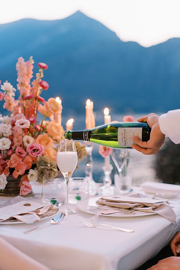 Reception tablescape wedding table setting with champagne bottle and flute, taper candles, and pastel florals on an outdoor terrace by a lake with mountain views at dusk