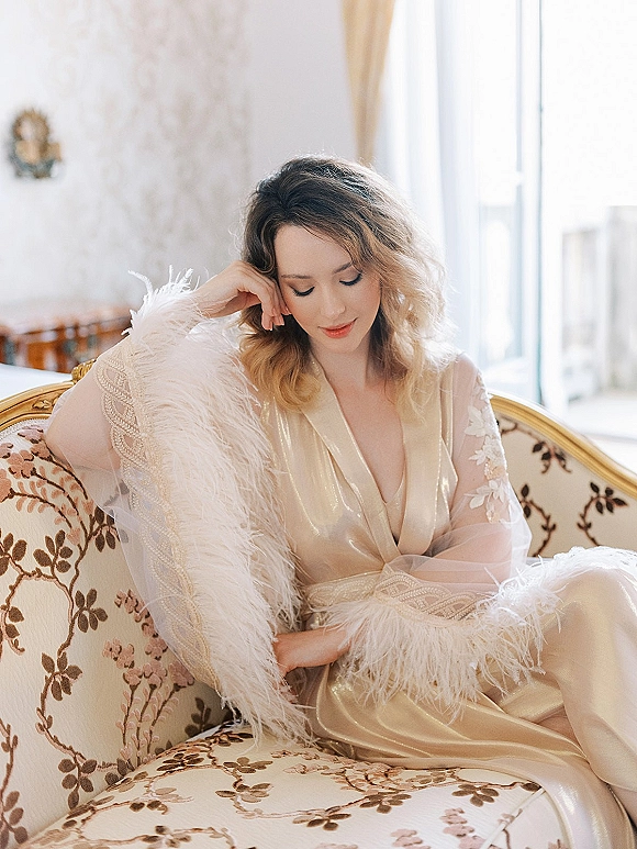 Bridal boudoir portrait of a bride in satin robe with feather trim, posed on a vintage floral sofa in soft window light