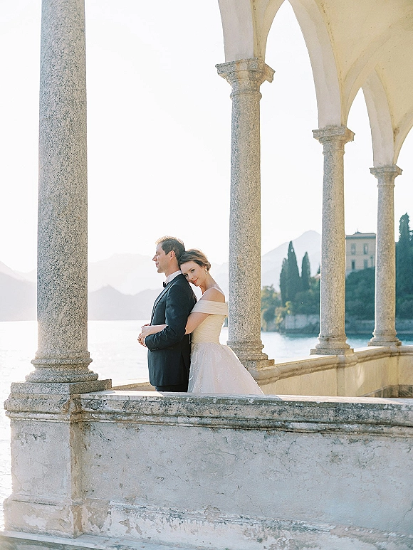 Couple portrait of bride and groom embrace in tuxedo and off-the-shoulder wedding dress under stone arches with lake and mountains beyond