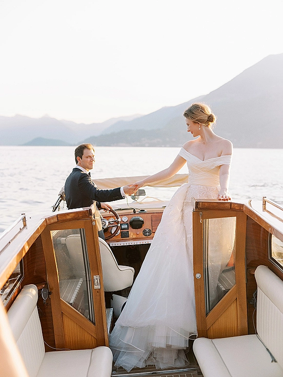 Couple portrait of bride and groom on boat, holding hands in wedding dress and tuxedo on a wooden motorboat with mountains beyond the lake
