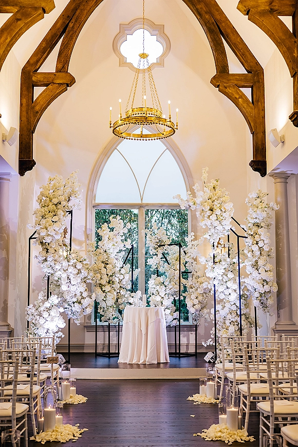 Ceremony setup with white floral pillars and a candle-lined aisle leading to a floral arch under a chandelier in an indoor chapel