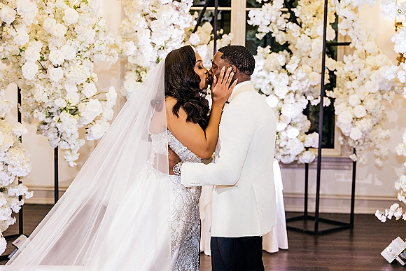 Wedding kiss portrait of bride and groom kissing, her hand on his face, with bridal veil and white floral backdrop in ballroom