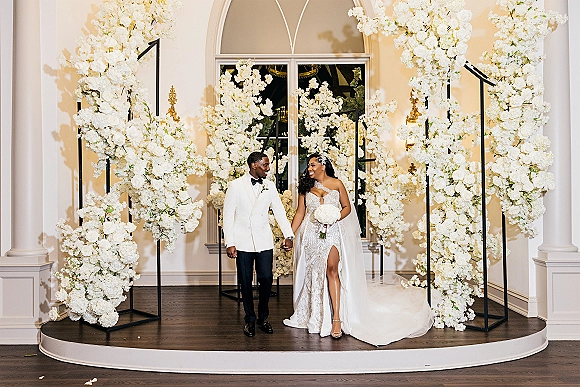 Couple portrait of bride and groom holding hands, she in strapless lace gown with veil and bouquet, under white floral arch indoors