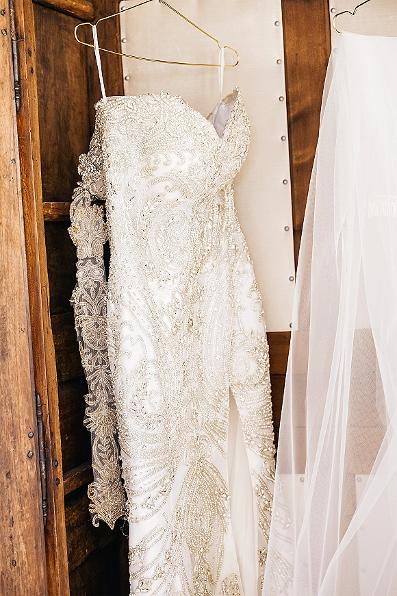 Wedding dress in a beaded wedding dress style hanging on a gold wire hanger, veil draped beside it against a wooden wardrobe backdrop