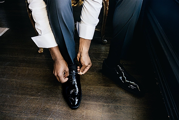 Groom getting ready, tying black patent wedding shoes with navy suit pants and cuffed white shirt on a wood floor by a chair
