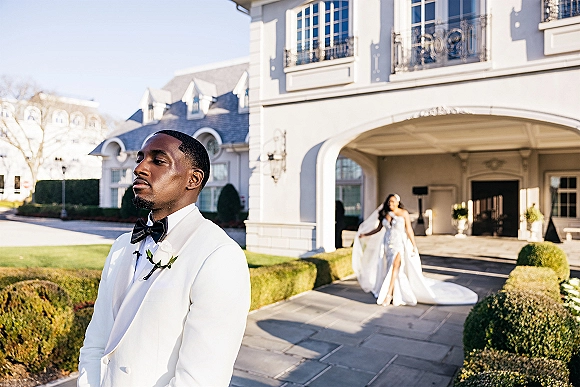 Wedding first look as groom in a white tuxedo waits under a mansion archway while the bride approaches in a veil and long train