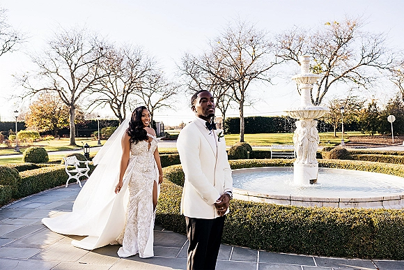 First look moment as bride in lace gown and long veil approaches groom in white tuxedo by a courtyard fountain and hedges