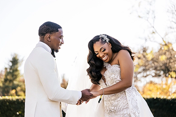 Couple portrait of bride and groom holding hands, laughing in profile; bride in strapless beaded dress with cathedral veil amid trees.