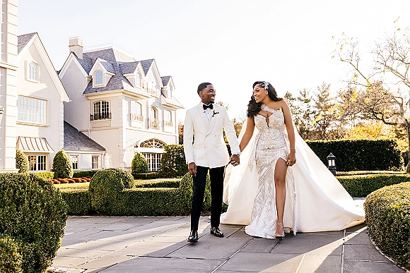 Couple portrait of bride and groom holding hands, bride in beaded gown with cathedral veil, strolling past a mansion and hedges.
