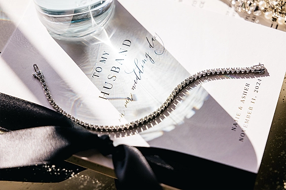 Invitation suite wedding invitation flat lay with envelope tied in black satin ribbon, styled with crystal bracelet, earrings, and perfume on light tabletop