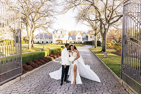 Couple portrait of bride and groom embrace, her veil train and bouquet visible by wrought iron gates at a grand estate garden drive