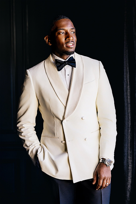 Groom portrait in an ivory tuxedo jacket and black bow tie, wearing cufflinks, watch, and wedding band against a dark curtain backdrop