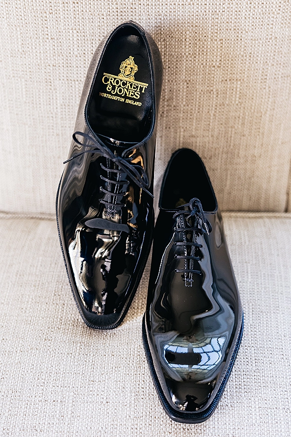 Groom shoes, black patent dress shoes with laces and gold logo print resting on beige textured fabric upholstery