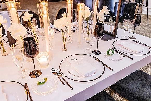 Reception tablescape with wedding table setting of white linens, taper candles, hurricane glass, white florals, and black goblets on a long table