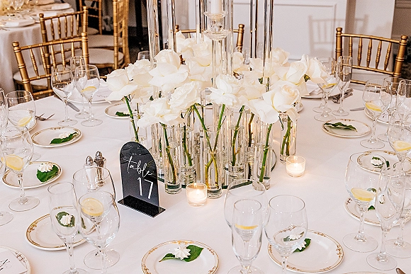 Reception tablescape with a white rose centerpiece in cylinder vases, tall taper candles, and white-and-gold place settings on a round table