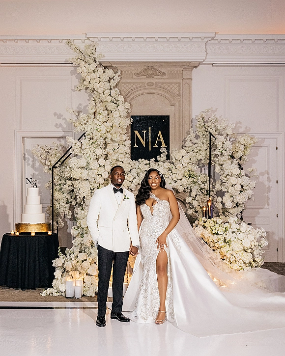 Couple portrait of bride and groom holding hands, her cathedral veil and his white tuxedo by a floral fireplace with candles and cake table