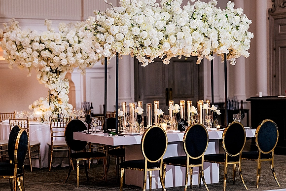 Reception tablescape with head table decor under a white floral canopy, taper candles and glassware, framed by a floral arch in a ballroom