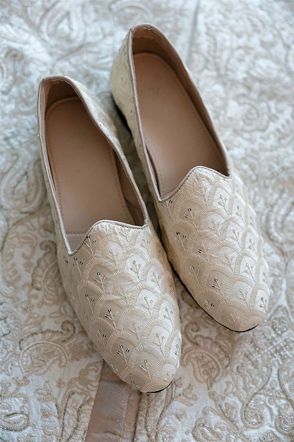 Wedding shoes in ivory bridal flats with metallic thread embroidery, resting on patterned textile and neutral bedding in a flat lay