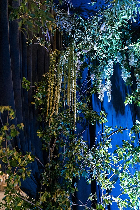 Wedding backdrop florals with a greenery wedding backdrop, white blooms and cascading garlands over draped fabric against a blue curtain with moody lighting