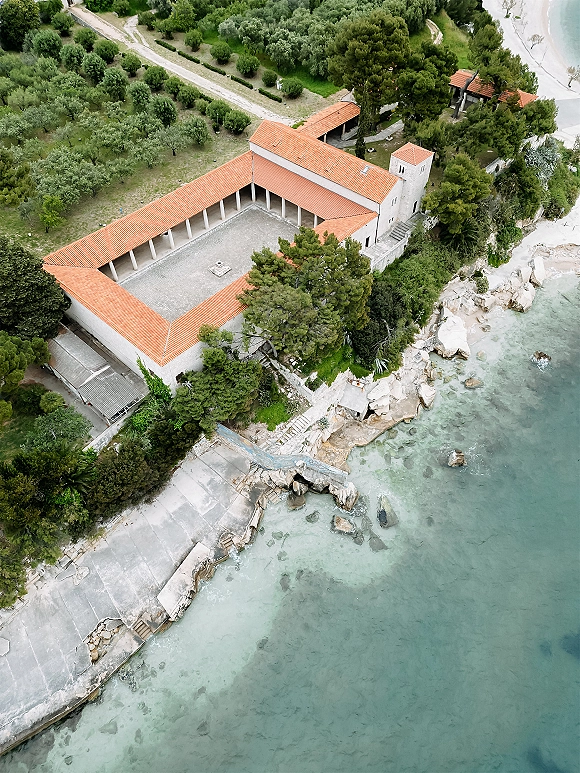 Wedding venue aerial view of a coastal wedding venue with terracotta roof stone villa, courtyard and tower beside turquoise ocean and rocky shore