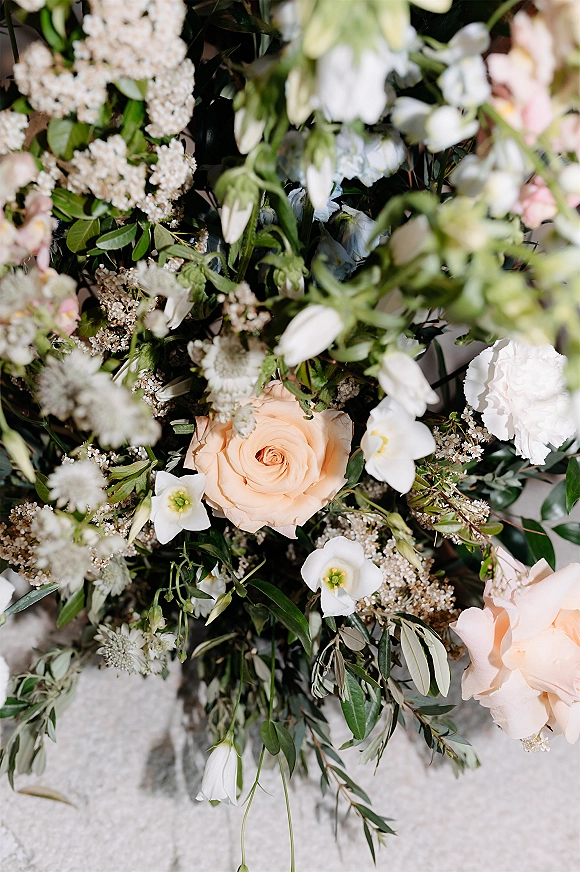Bridal bouquet with peach rose bouquet tones, white blooms and greenery resting on a light stone surface with soft foliage accents