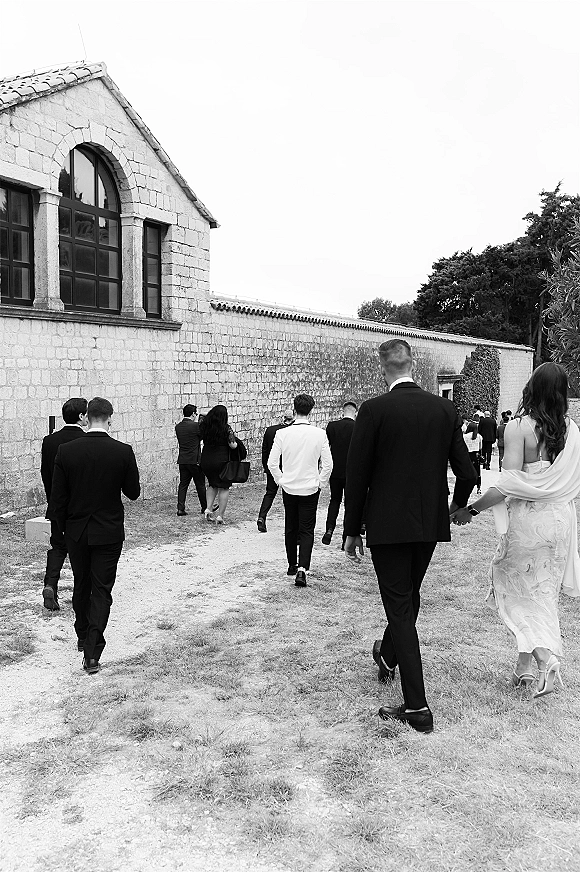 Wedding guests walking together in suits and dresses, one in a shawl, along a gravel path beside a historic stone building and trees