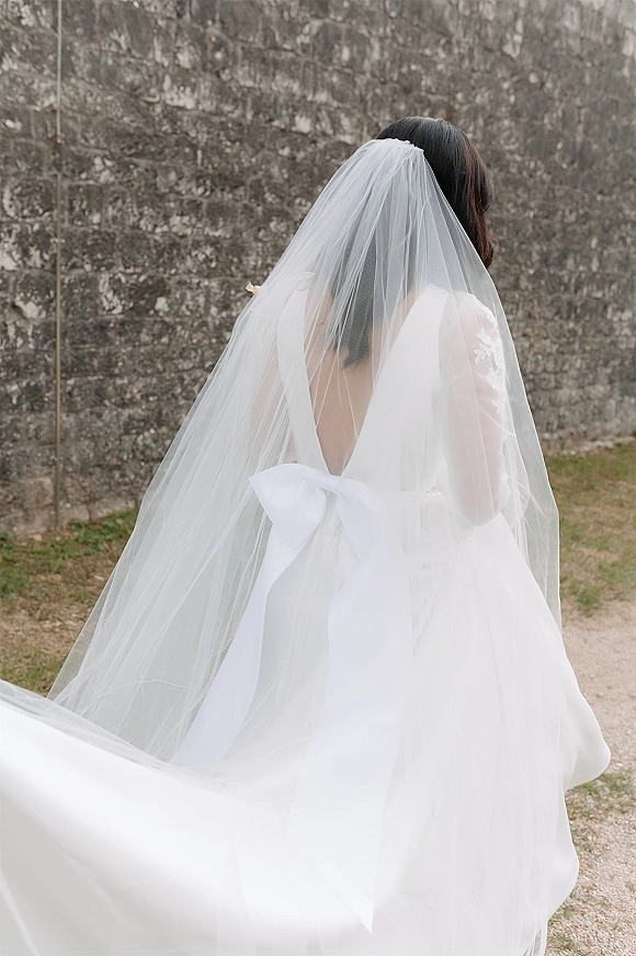 Bridal portrait from behind with long veil flowing over a white gown with oversized bow, walking along a gravel path by a stone wall